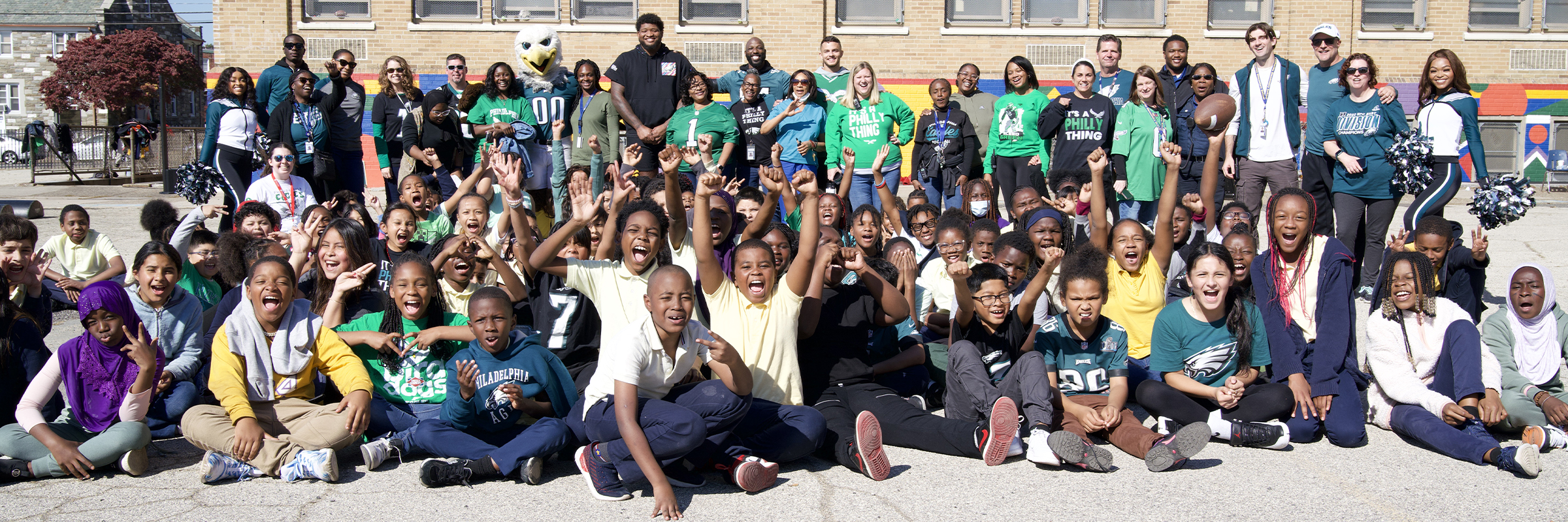 A large group of Joseph W. Catharine School students and staff cheer and smile during an outdoor celebration featuring Philadelphia Eagles cheerleaders, the team mascot, and community guests, showcasing school spirit and partnership.