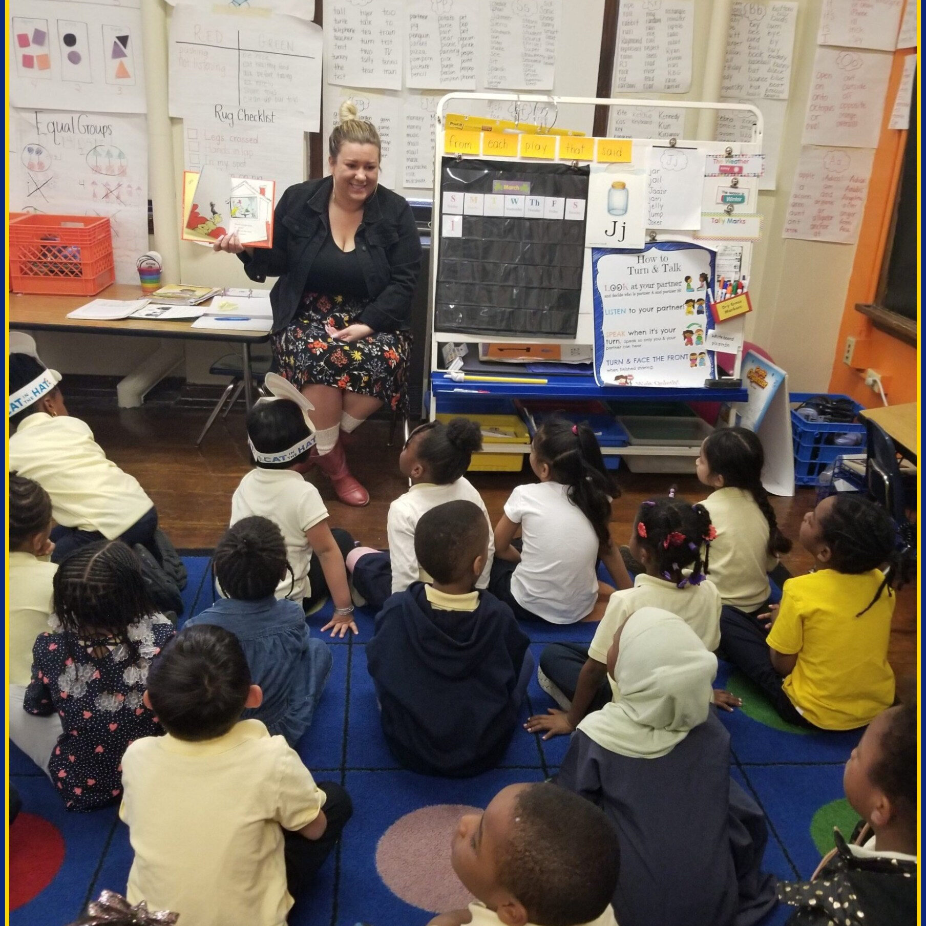 Image Border Editor: https://www.tuxpi.com/photo-effects/borders A teacher reads a picture book to a group of young students seated on a colorful classroom rug. The children listen attentively, surrounded by educational posters and charts focused on reading, writing, and vocabulary at Joseph W. Catharine Elementary.
