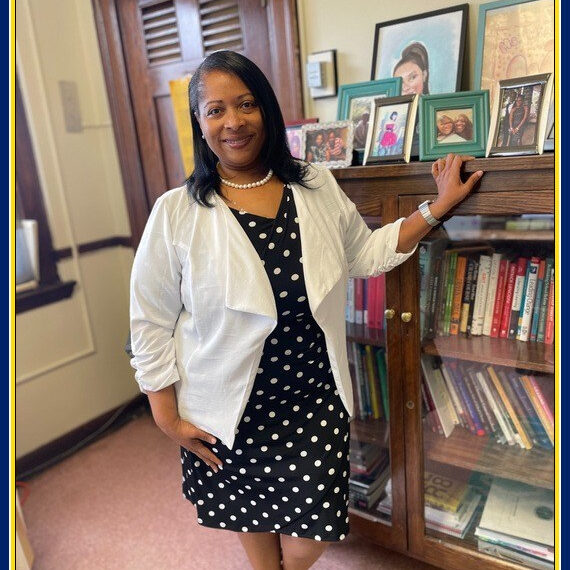 Principal Wilmer in Her Office Principal Wilmer stands smiling in her office, wearing a black polka dot dress with a white blazer. She is leaning against a wooden bookcase filled with books and family photos, with framed pictures displayed on top.